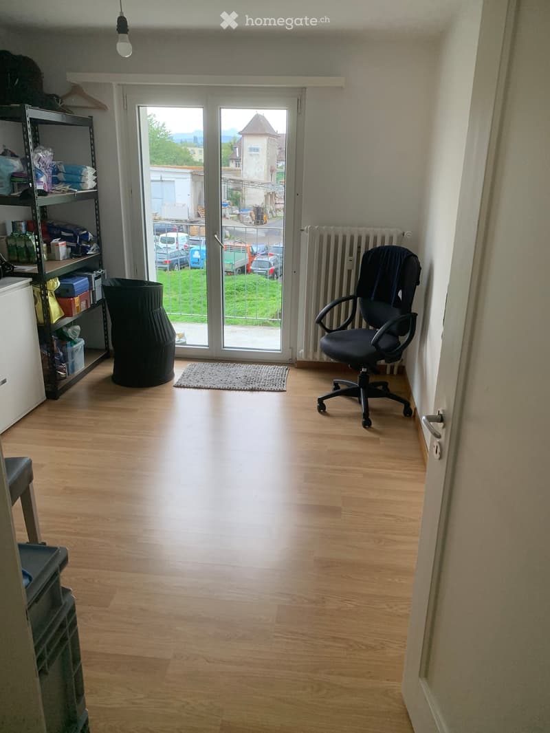 A small, well-lit room with wooden flooring, a large window with a view of the outside, a black office chair, a shelf with various items, and a radiator.