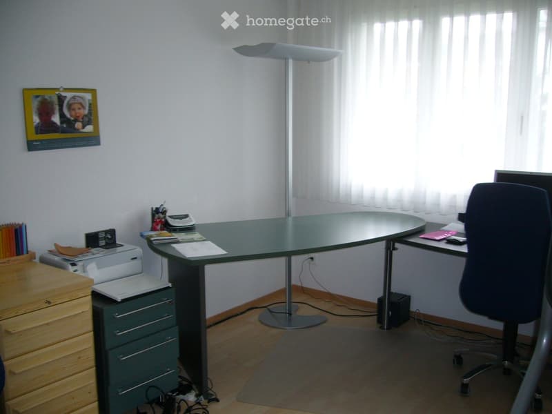 A well-lit office space with a large glass desk, ergonomic chair, printer, floor lamp, and wooden cabinet.