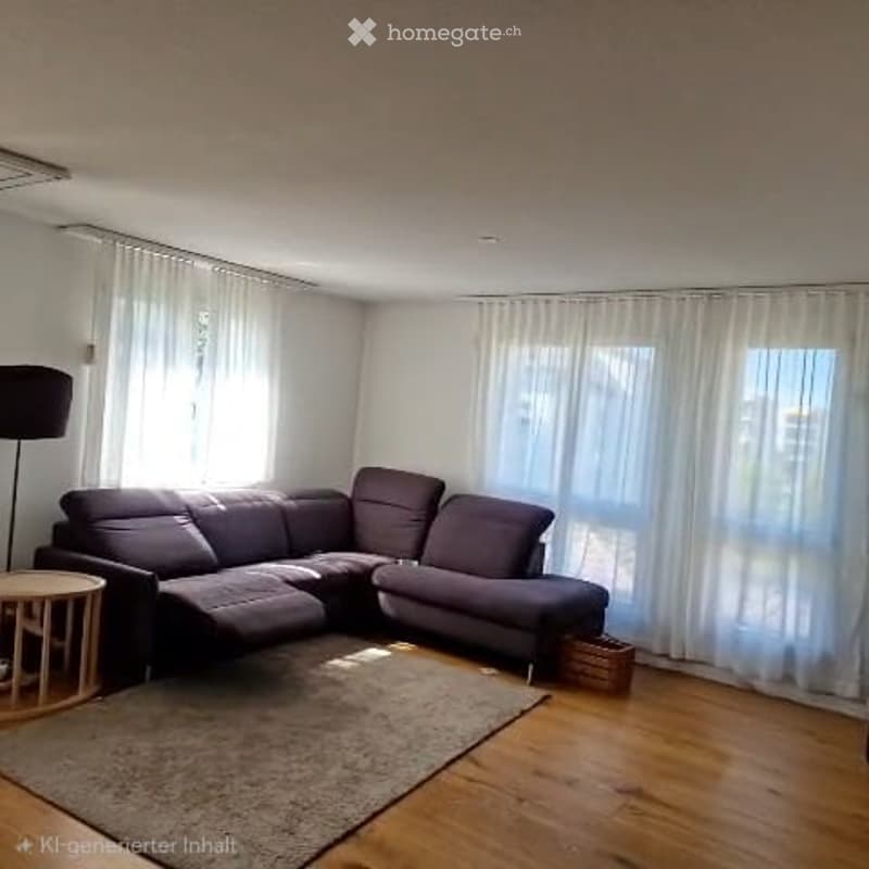 Modern living room with a dark grey sectional sofa, a small round wooden coffee table, a floor lamp, and a large area rug on a wooden floor. Large windows with white curtains allow natural light to fill the space.