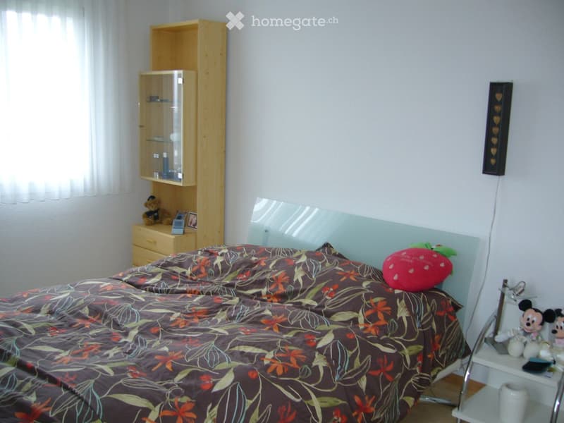 A well-lit bedroom with a large bed covered in a floral-patterned duvet, a nightstand with a lamp, a mirror, a wooden shelf with decorative items, and a small table with a white mug and Mickey Mouse toys.