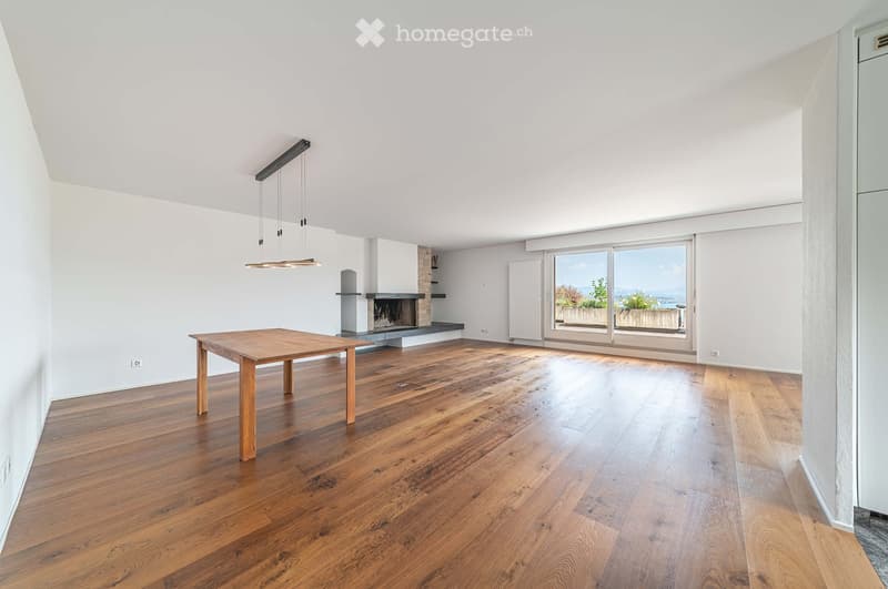 Bright, open living space with wooden flooring, a fireplace, a wooden table, and large windows offering a scenic view.