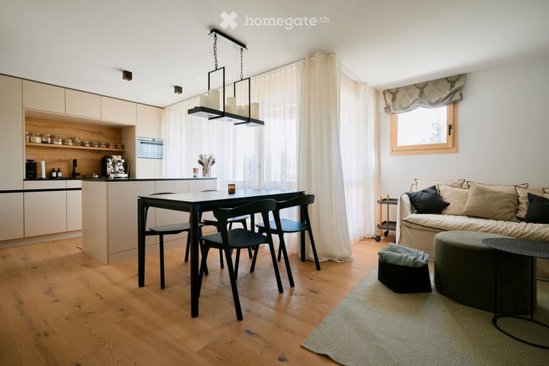 Open-plan living and dining area with modern furniture, including a sofa, coffee table, dining table with chairs, and kitchen cabinets. The space features wooden flooring, large windows with curtains, and a hanging light fixture.