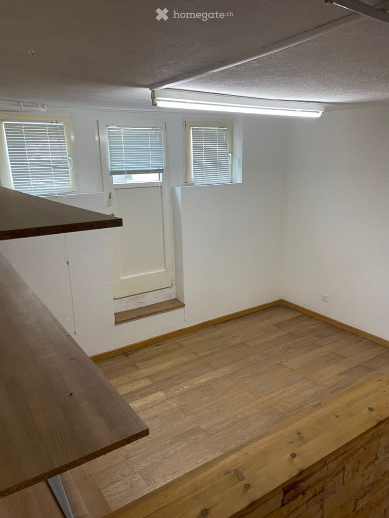 Empty room with wooden flooring, white walls, three windows with blinds, and a small wooden bench near the door.