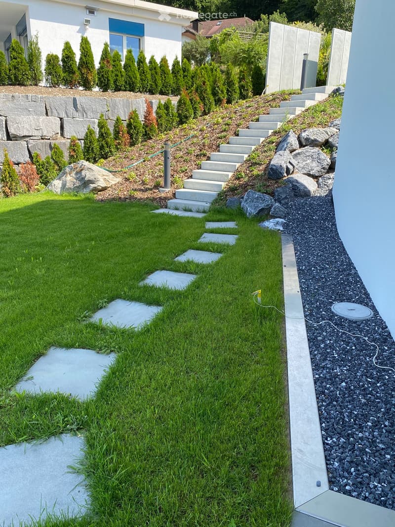 Well-maintained garden with a lawn, concrete steps, gravel pathway, and surrounding trees and shrubs.