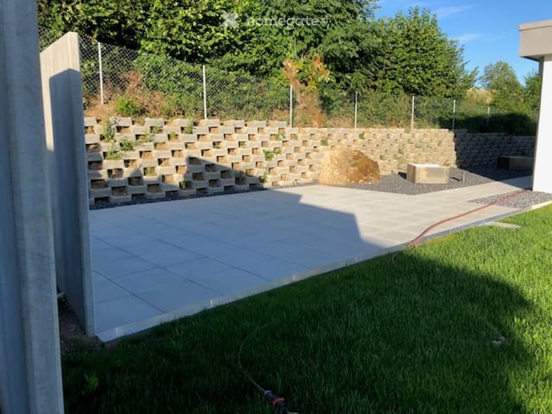 A modern outdoor patio area with a concrete floor, a stone retaining wall, and a grassy lawn. The area is enclosed by a metal fence and features a large stone block and a small concrete bench.