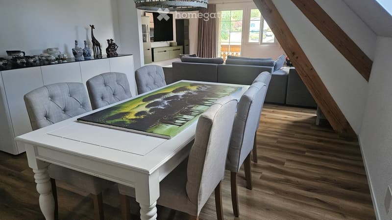 Dining area with a white wooden table, four grey upholstered chairs, and a large artwork on the table. A grey sectional sofa is visible in the background, along with a television mounted on the wall and decorative vases on a white cabinet.