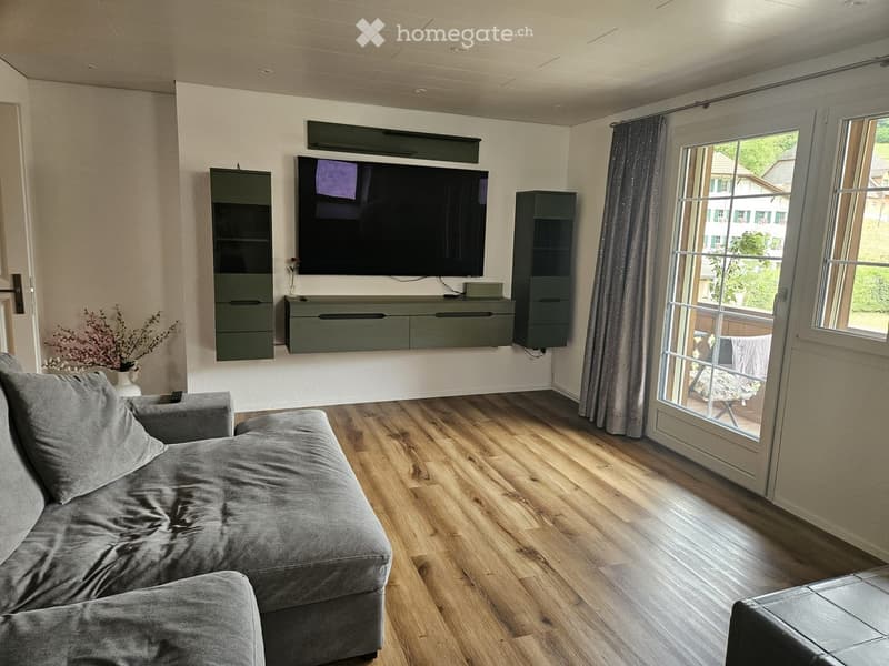 Modern living room with a grey sectional sofa, large flat-screen TV mounted on the wall, grey cabinets, and a vase with dried flowers on a side table. Large windows with curtains lead to a balcony.