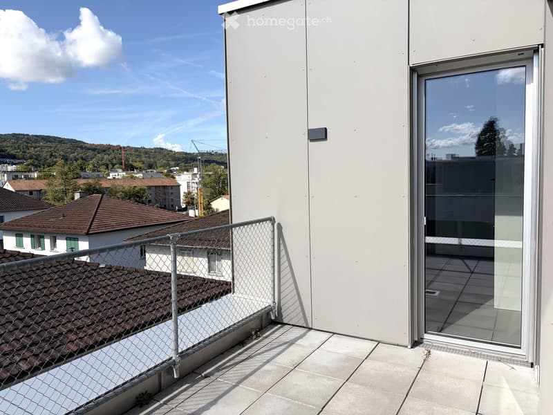 Modern rooftop terrace with a glass door, metal railing, and tiled floor, overlooking a cityscape with houses and trees.