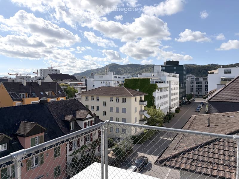 View from a balcony showing a cityscape with various buildings, rooftops, and a partly cloudy sky.