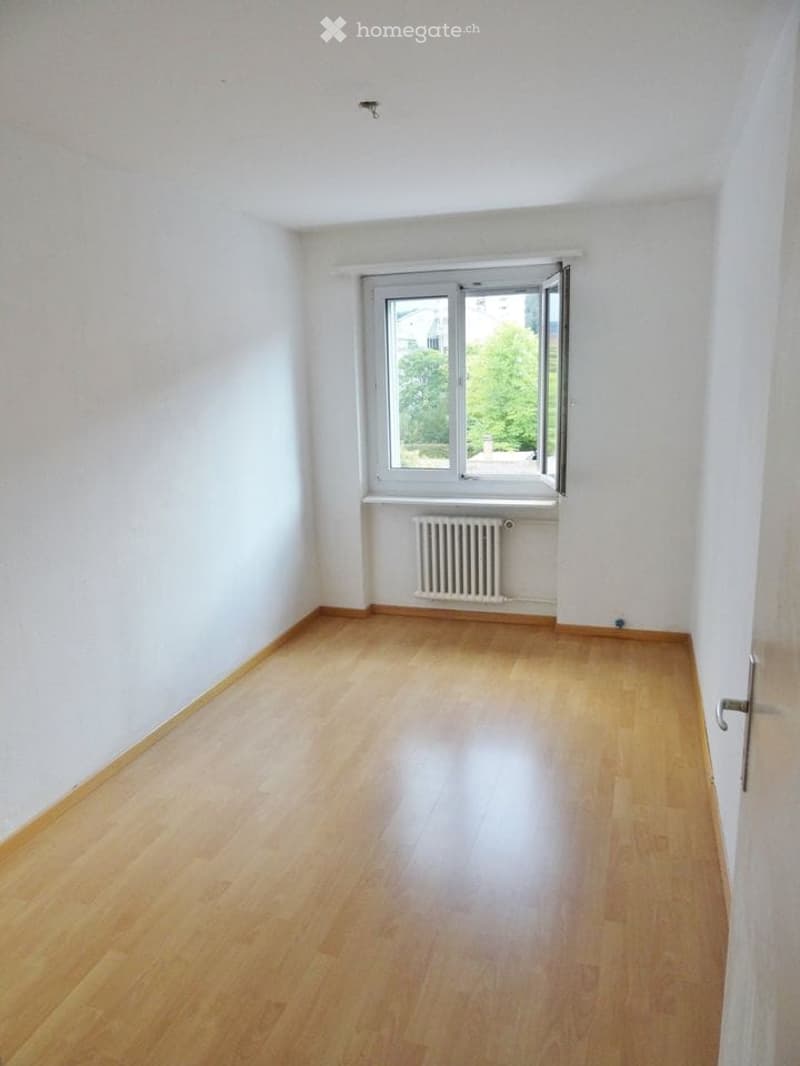 Empty room with light wooden flooring, white walls, and a window with a radiator below it.