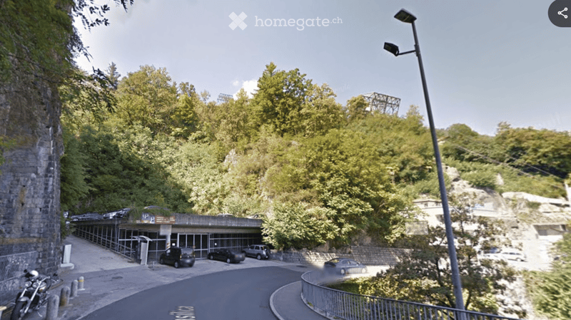 A parking area with several cars parked, surrounded by greenery and a stone wall, with a modern building entrance visible.