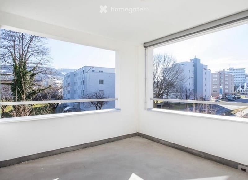 Empty room with large windows offering a view of the city and trees, white walls, and a concrete floor.