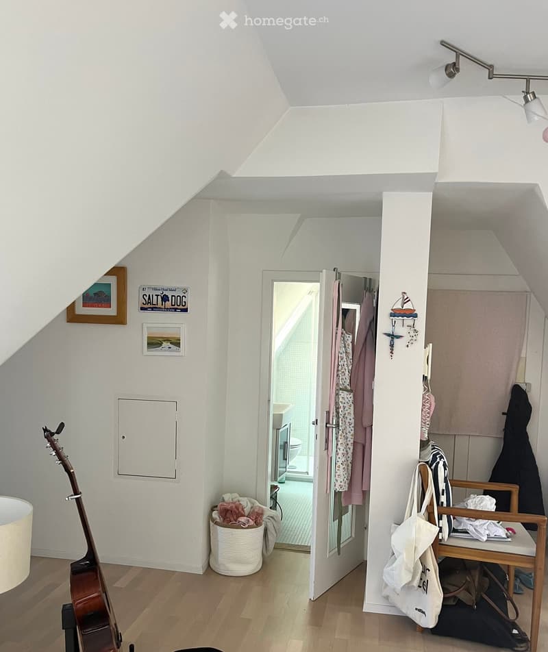 A small, well-lit room with wooden flooring, a slanted ceiling, and a guitar leaning against the wall. A stool with a handbag and other items sits near the door, which leads to a bathroom. A basket with clothes is placed near the door, and two picture fra