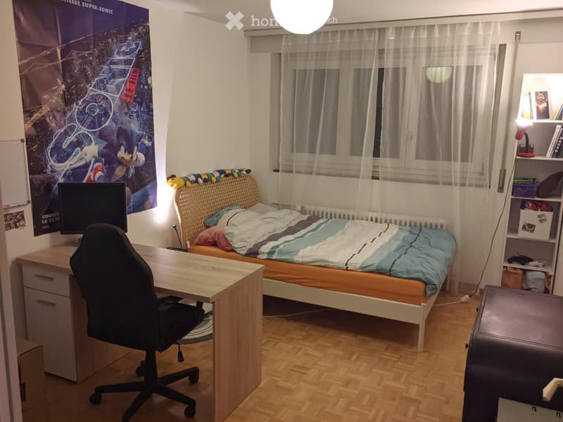 A tidy bedroom with a single bed, a desk with a chair, a large window with sheer curtains, and a shelf with books and decorative items.