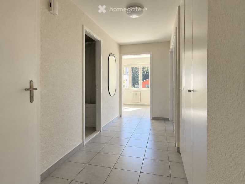 Clean, modern hallway with tiled flooring, white walls, and a round mirror on one wall. Doors lead to adjacent rooms, and natural light streams in from an open doorway at the end.