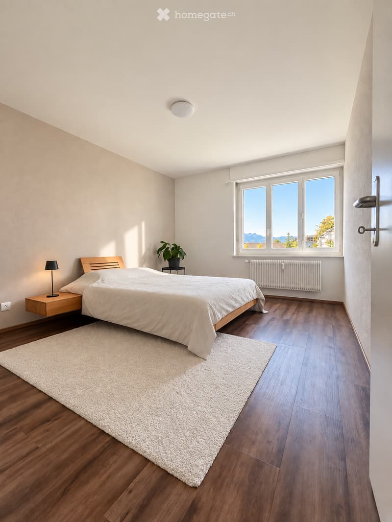 Modern bedroom with wooden flooring, large window offering a scenic view, a neatly made bed with white bedding, a nightstand with a lamp, and a small potted plant on a side table.