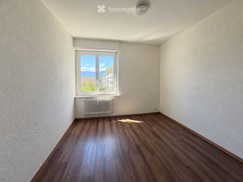 Empty room with wooden flooring, white walls, a window with a view, and a radiator below the window.