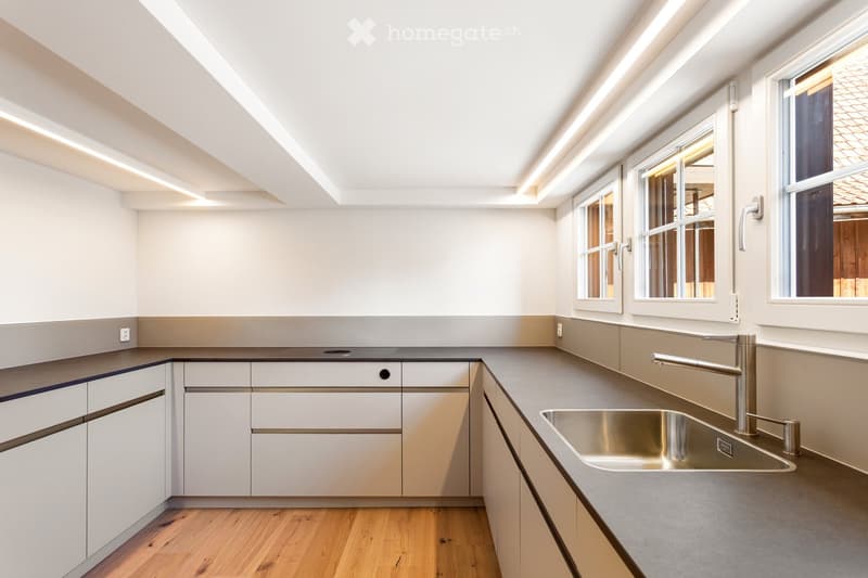 Modern kitchen with white cabinets, stainless steel sink, and sleek faucet. Natural light from large windows illuminates the space.