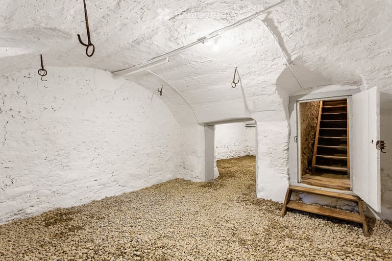 Empty room with white stone walls and a gravel floor, featuring a wooden staircase leading to an adjacent space.