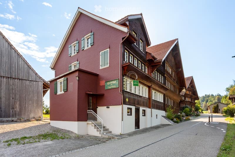 Traditional two-story building with red brick facade and wooden elements, featuring multiple windows with shutters, a staircase leading to the entrance, and a signboard above the door.