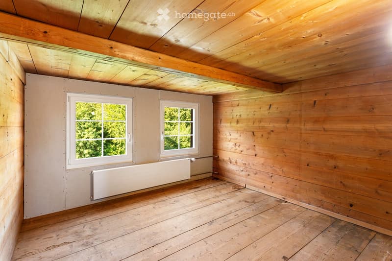 Empty wooden room with exposed beams, wooden floor, two windows, and a radiator.
