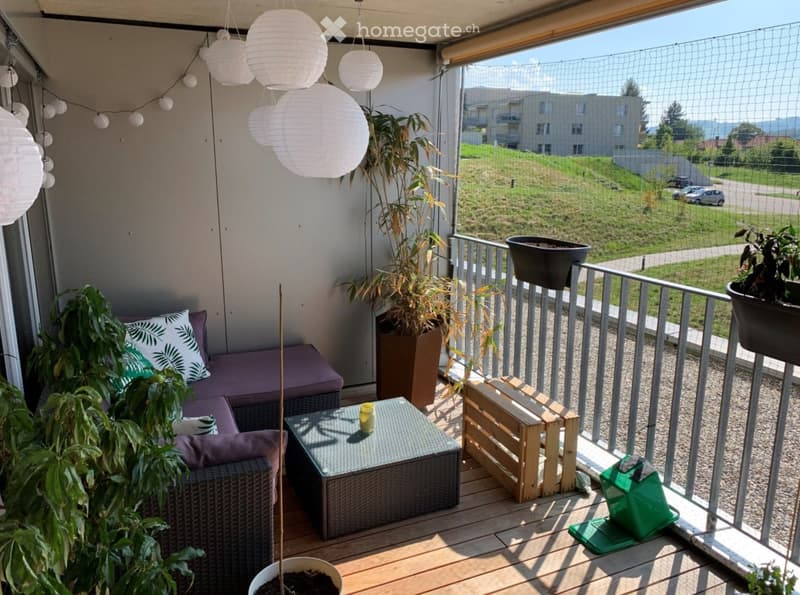 Balcony with wooden floor, sofa, small table, potted plants, and a view of a green area with buildings in the distance.