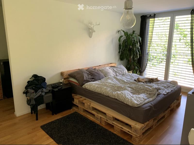 Cozy bedroom with a wooden pallet bed, nightstand, cat, and a large window with blinds.