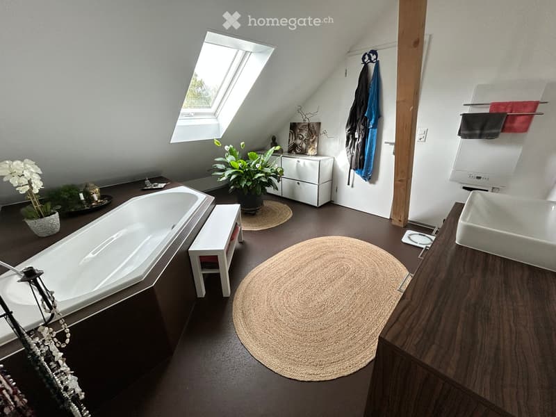 Modern bathroom with a freestanding bathtub, sink, small white stool, potted plant, and a round woven rug on the dark floor. Features a skylight window and a towel rack with hanging towels.