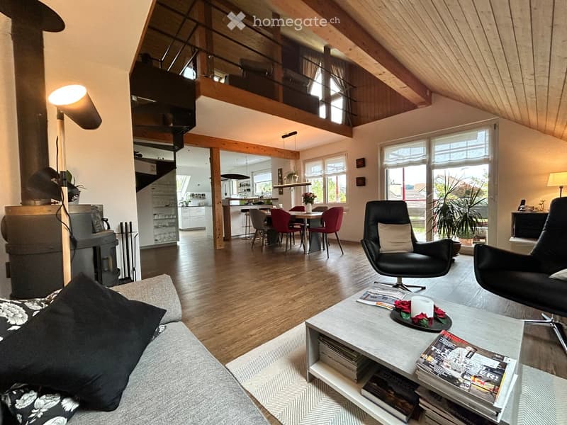 Open-plan living and dining area with wooden floors, exposed wooden beams, large windows, a sofa, coffee table, armchairs, dining table with chairs, a fireplace, and a staircase leading to an upper level.