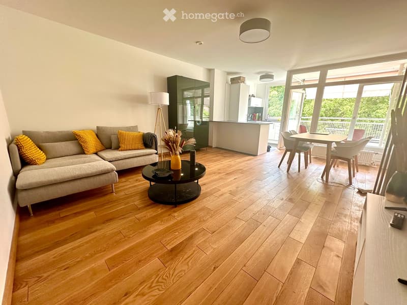 Modern living room with wooden flooring, grey sectional sofa with yellow cushions, black coffee table, floor lamp, dining area with table and chairs, and a balcony view.