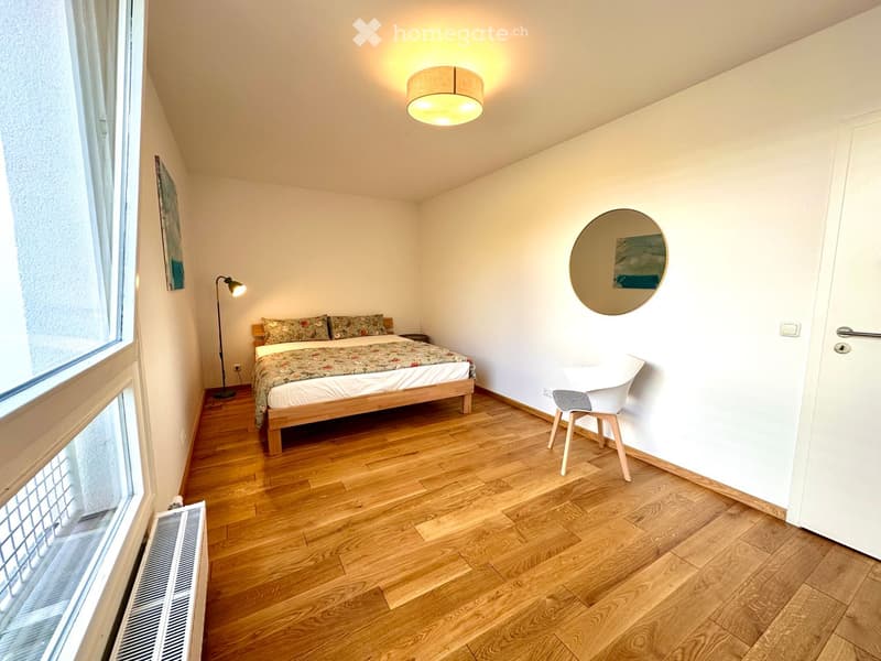 Cozy bedroom with wooden flooring, a double bed with patterned bedding, a nightstand, a white chair, a round mirror, and a floor lamp.
