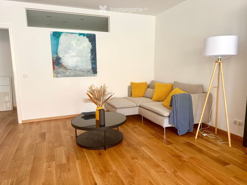 Modern living room with light wooden flooring, a grey sectional sofa with yellow cushions, a round black coffee table, a tall floor lamp, and a decorative painting on the wall.