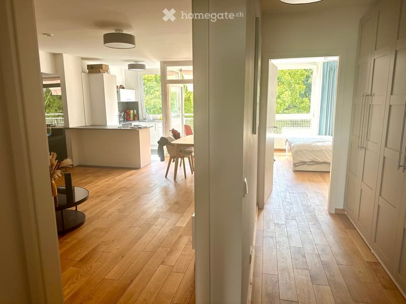 The image shows a modern, well-lit interior with a kitchen area, a dining area, and a bedroom. The kitchen features white cabinets, a sink, and a countertop. The dining area includes a table with chairs. The bedroom has a bed, a wardrobe, and a window wit