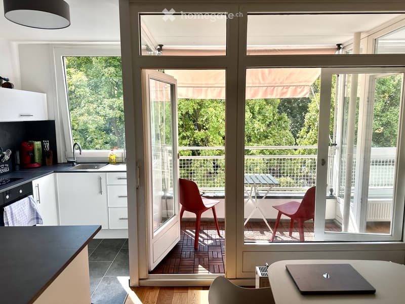 Modern kitchen with white cabinets, black countertops, and a red chair near the open sliding doors leading to a balcony with a small table and chairs.