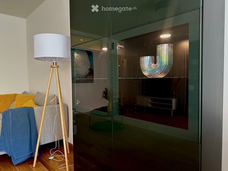 Modern living room with a grey sofa, yellow pillows, a blue throw, a wooden floor lamp, and a glass partition reflecting a TV stand and decorative lamp.