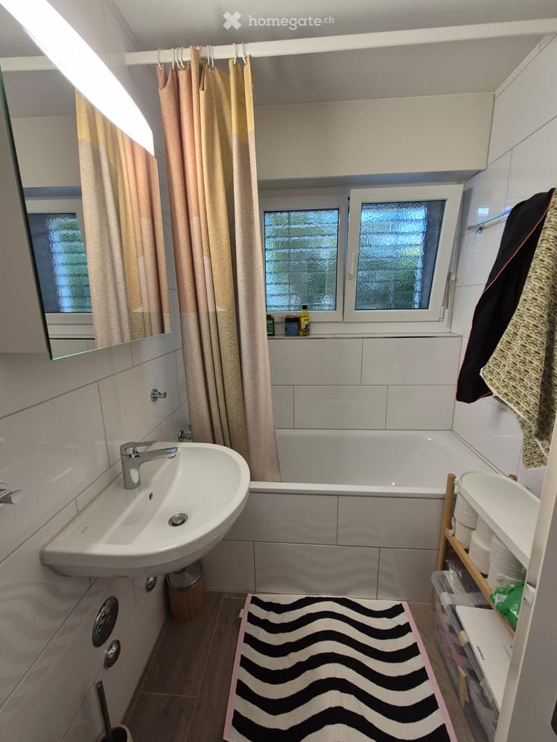 Modern bathroom with a white sink, bathtub, mirror, and tiled walls. Includes a towel rack with towels, a small shelf with toiletries, and a patterned rug on the floor.