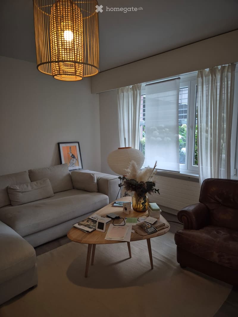 Modern living room with a grey sectional sofa, a round wooden coffee table, a brown leather armchair, a large pendant light, and a vase with dried flowers on the coffee table.