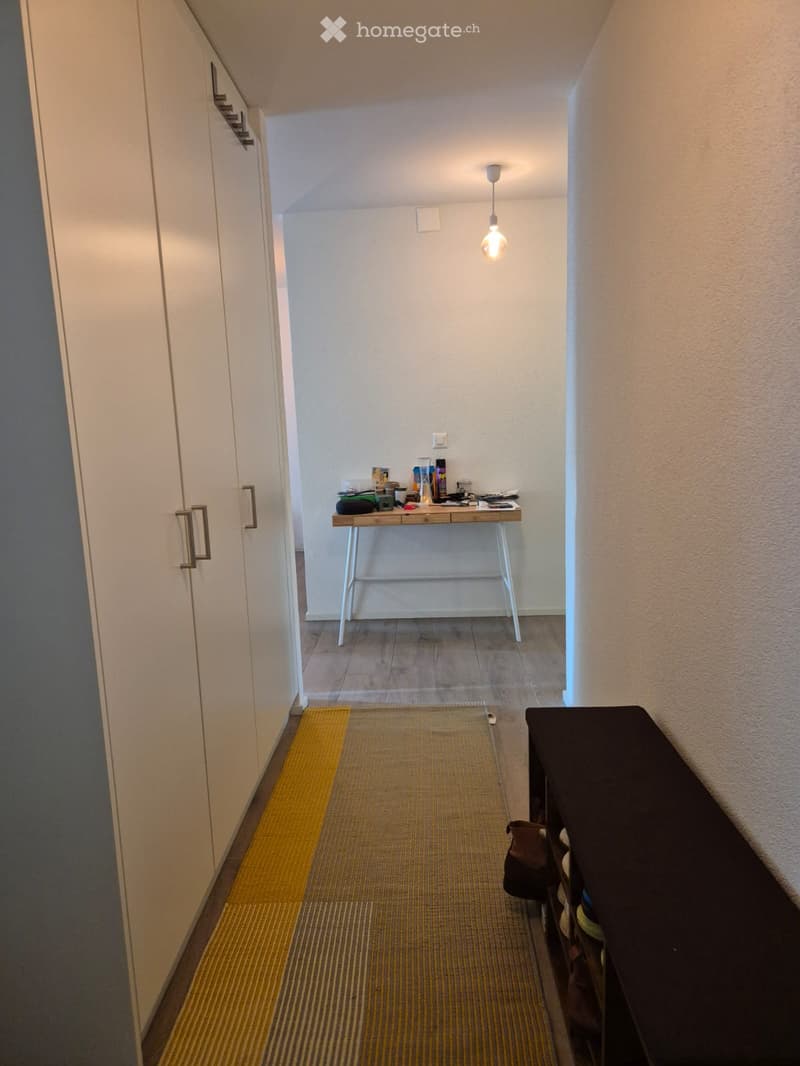A narrow hallway with white walls, a hanging light fixture, a small table with various items, and a yellow runner on the floor.