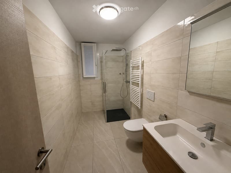 Modern bathroom with beige tiles, a glass-enclosed shower, a white toilet, a sink with a modern faucet, and a large mirror above the sink.