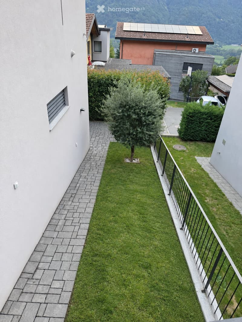A small, well-maintained garden area with a neatly trimmed lawn, a small tree, and a paved pathway leading to the building entrance. The garden is bordered by a metal railing and is surrounded by neighboring houses and greenery.