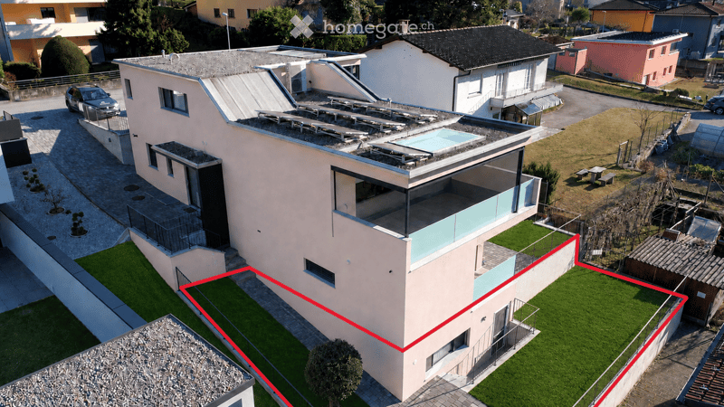 Modern two-story house with a flat roof, large windows, and a swimming pool on the roof. The house has a light-colored facade and is surrounded by a well-maintained garden with a driveway and a parked car.