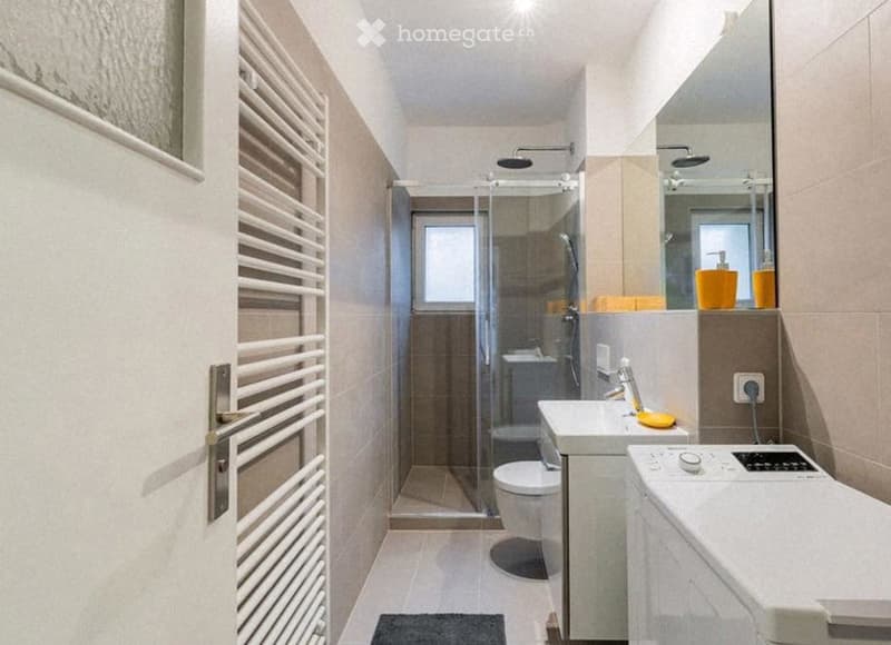 Modern bathroom with a glass-enclosed shower, toilet, sink, mirror, radiator, and washing machine. Features include a small window, modern fixtures, and a sleek design.