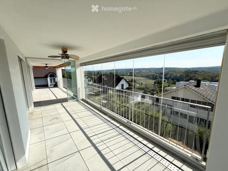 Spacious balcony with modern glass railing, tiled floor, and scenic view of surrounding houses and countryside.
