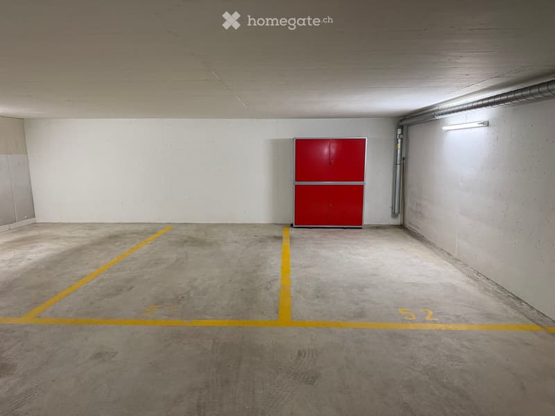 Empty underground garage with concrete floor, white walls, red storage cabinet, and yellow parking lines marked '51' and '52'.