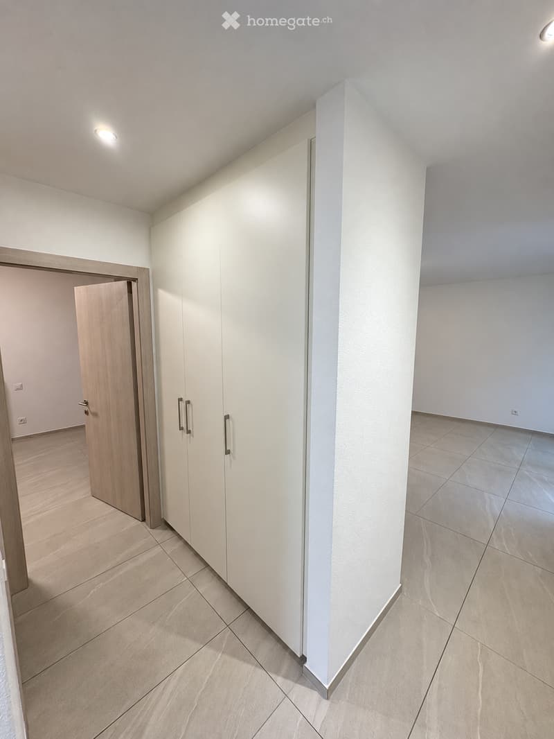 Empty room with light-colored tiled flooring, white walls, and built-in white cabinetry with silver handles. The room features recessed lighting and an open doorway leading to another space.