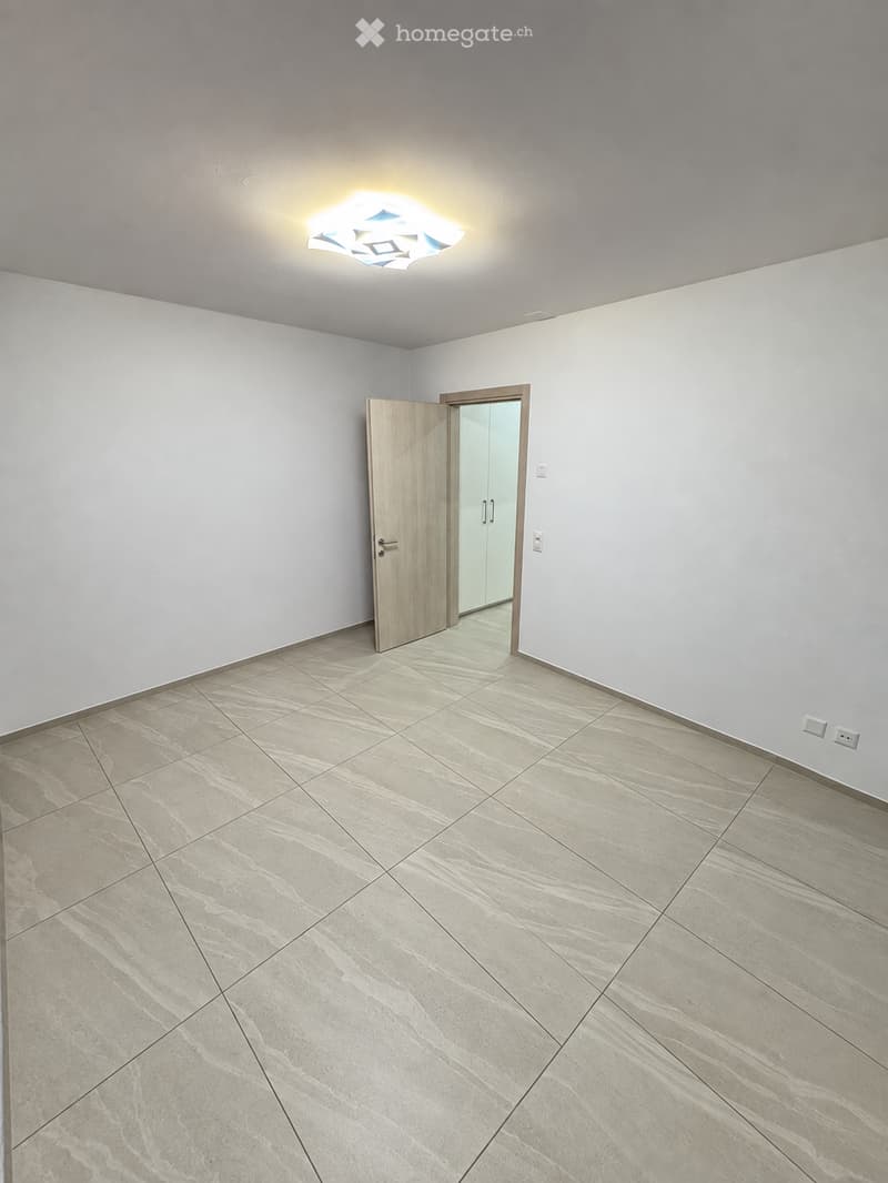 Empty room with light grey walls, beige tiled floor, and a single modern ceiling light fixture. A wooden door with a glass panel is open, revealing a glimpse of another room.