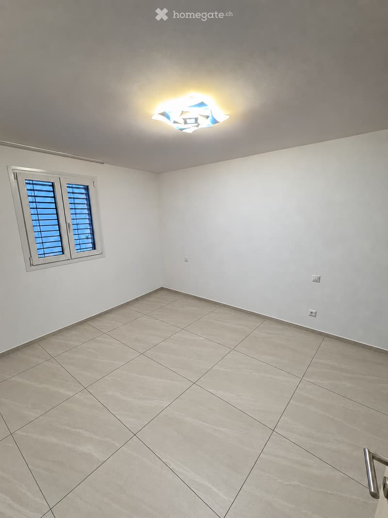 Empty room with light grey walls, beige tiled floor, and a single window with blinds. A modern ceiling light fixture is visible.