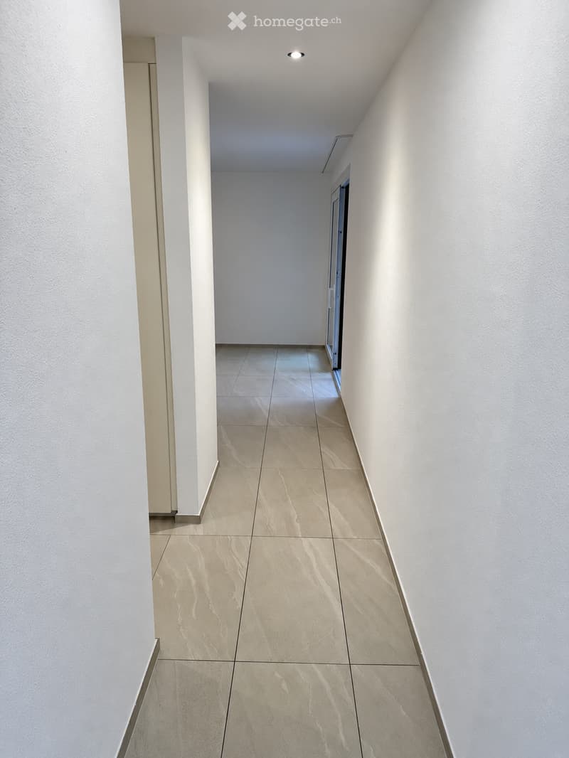 Modern hallway with beige tiled flooring, white walls, and recessed ceiling lighting. Features a partially visible door at the end.