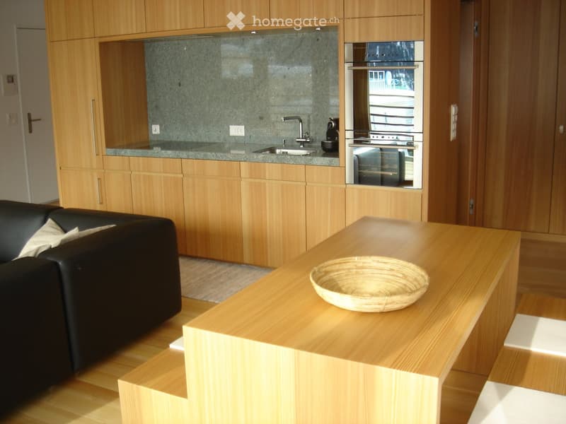 A modern, open-plan living and kitchen area with a black leather sofa, a wooden table with a woven basket, and a kitchen featuring wooden cabinets, a stainless steel oven, and a sink.