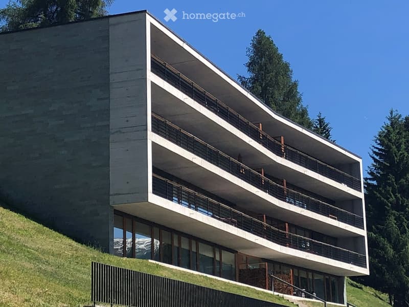 Modern multi-story building with balconies, large windows, and a raised ground floor, set against a backdrop of trees and mountains.